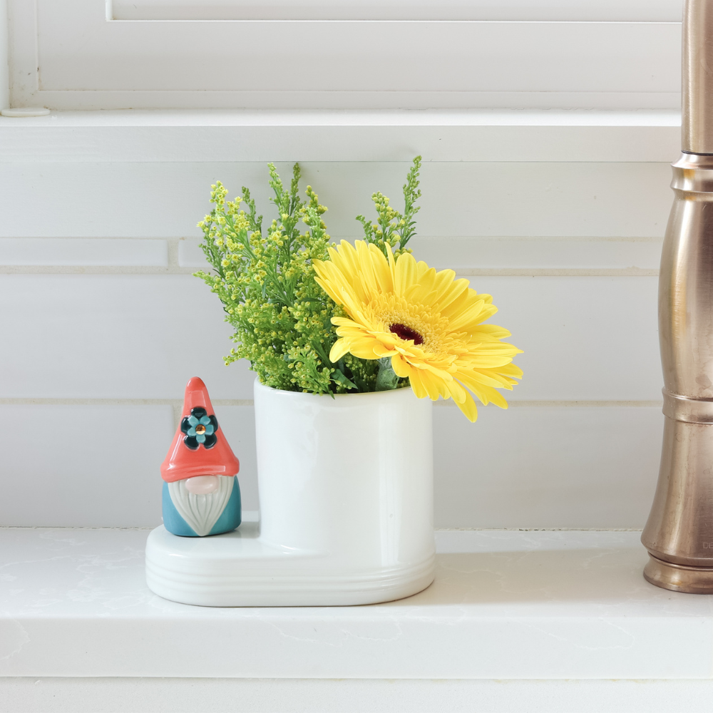 'oh gnome you didn't' mini of a gnome that is blue and orange with a blue flower on the hat on a cutie container filled with an yellow flower and on the kitchen counter next to the sink.