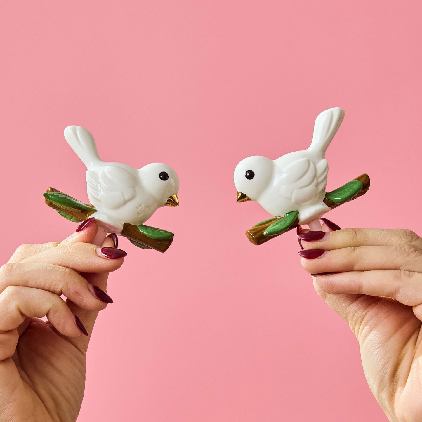 White ceramic bird minsi on a branch with a pink background and hands holding them up