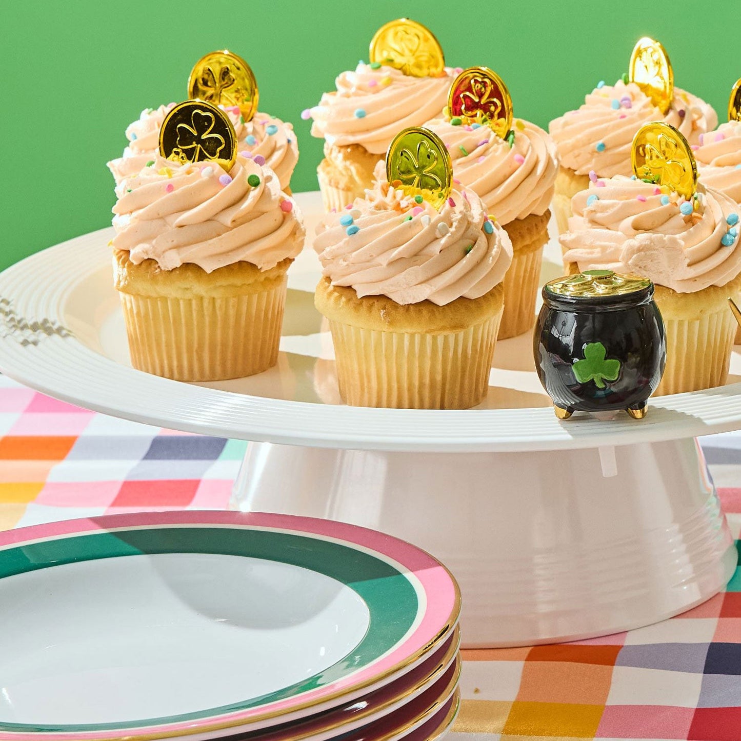 Black pot with a green clover mini on a white surface with cupcakes against a green background and plaid tablecloth
