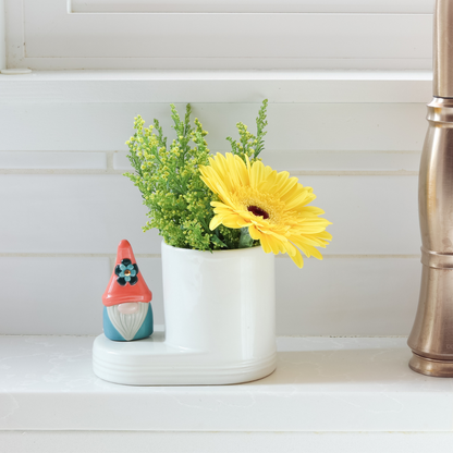 'oh gnome you didn't' mini of a gnome that is blue and orange with a blue flower on the hat on a cutie container filled with an yellow flower and on the kitchen counter next to the sink.