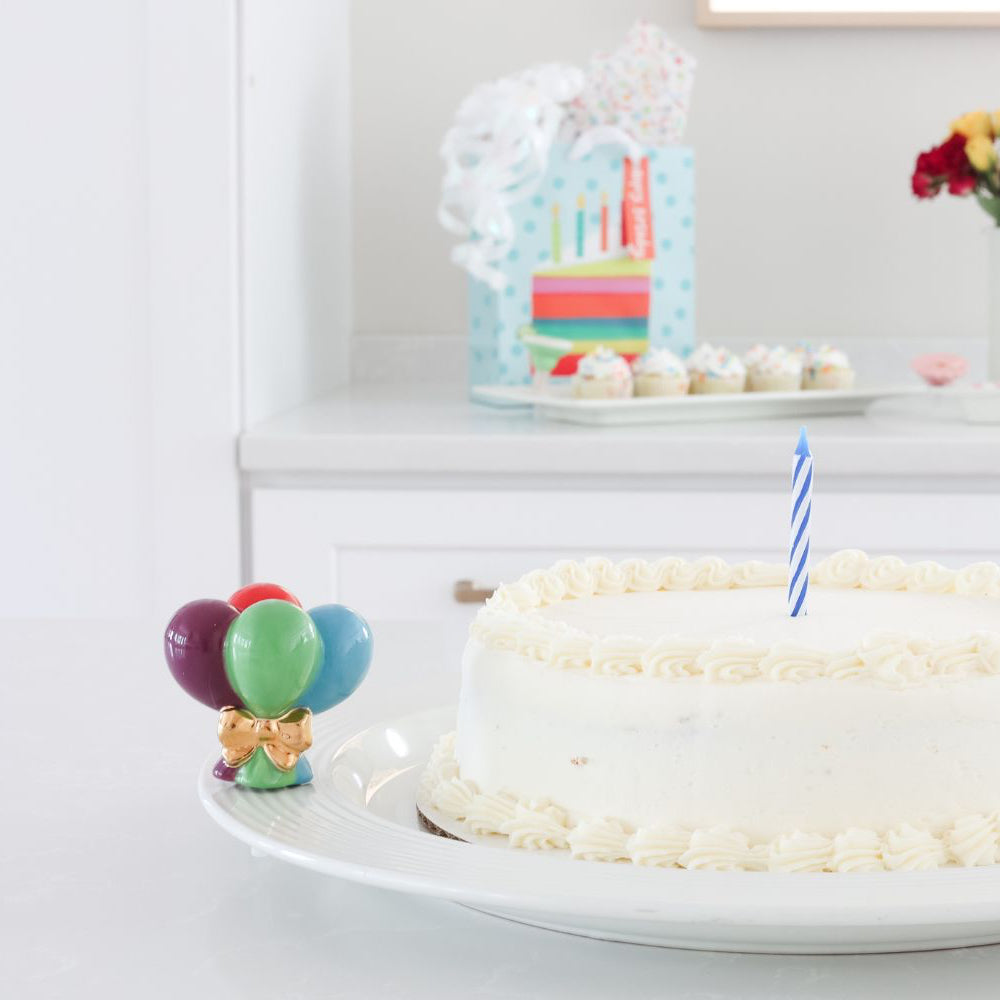 A ceramic mini four colorful balloons wrapped in  a gold bow on a round platter that holds a white birthday cake with one candle.