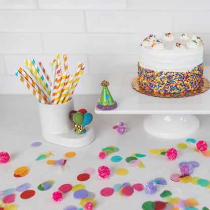 green, blue and purple celebration hat to commemorate 20 years of nora fleming on top of a white cake stand next to a cutie container filled with straws and topped with balloon mini.