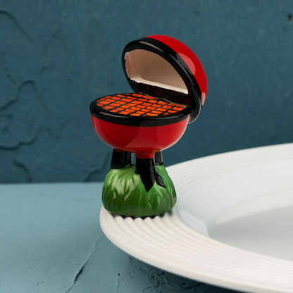 red barbecue grill mini on a green base with a white plate against a blue wall.
