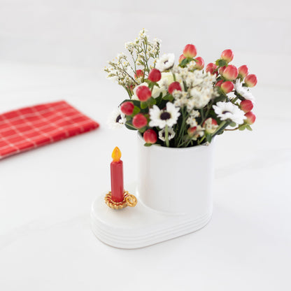 This image features a charming "Light the Way" mini candle holder, perfect for enhancing the festive spirit during the holiday season. The candle holder, with its vintage-inspired design, is depicted in a warm, glowing hue of gold, complementing the bright red candle it holds. The mini is sitting on a white cutie container that holds a holiday floral arrangement.